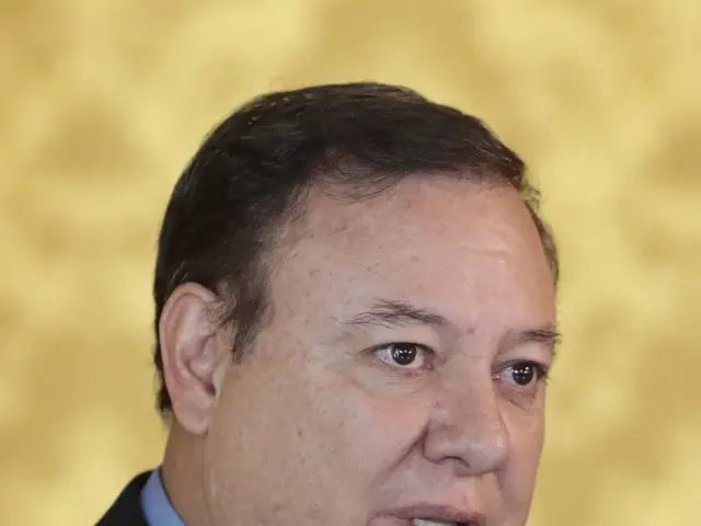 The image shows a man in a suit and tie speaking into a microphone in front of a blurred...