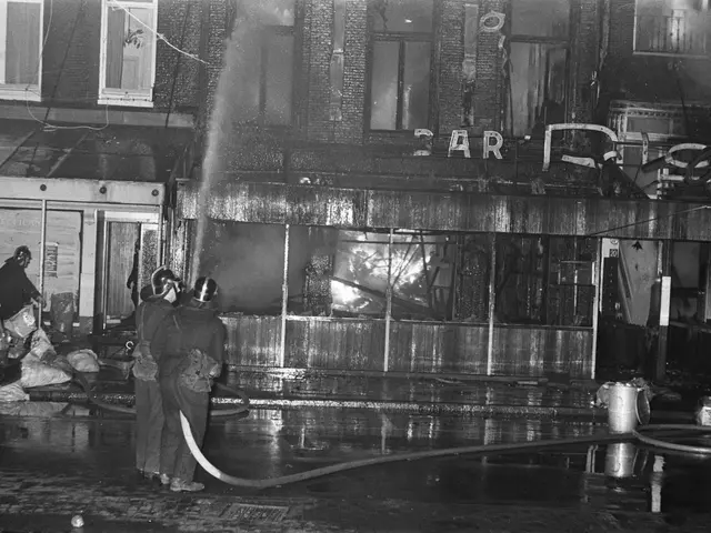 The image shows a black and white scene of firefighters working to put out a fire on the corner of...