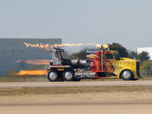 The image shows a truck with flames coming out of the back of it driving down a road surrounded by...