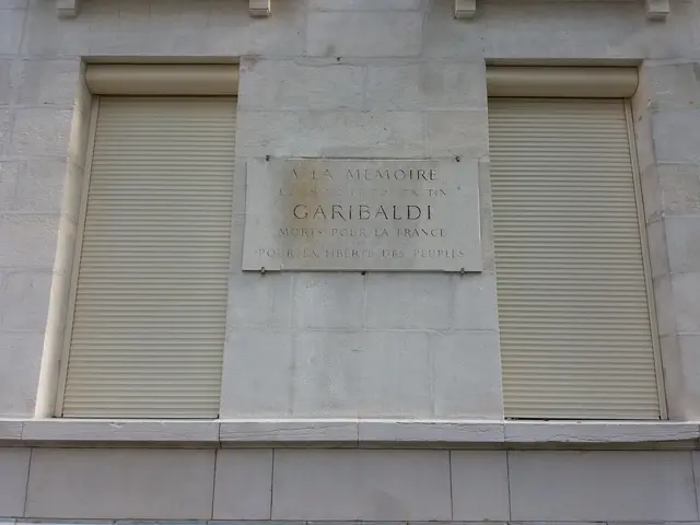 The image shows a building with two windows and shutters, and a plaque on the side of the building....