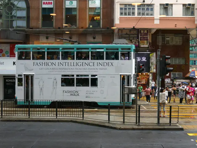 The image shows a double decker bus driving down a street lined with tall buildings. There are many...