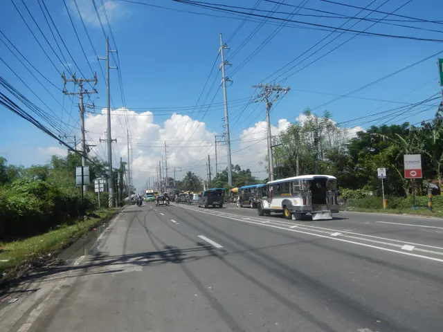 The image shows a city street with cars and buses driving down it, surrounded by electric poles...