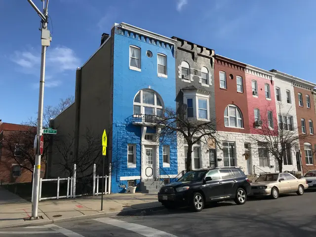 The image shows a row of colorful houses on the corner of a street, with cars parked on the side of...