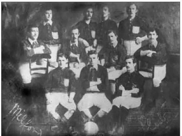 The image shows a black and white photo of a football team from the 1920s, with the players sitting...