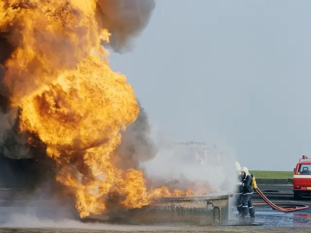 The image shows a fire truck engulfed in flames on the side of a road, with two people wearing...