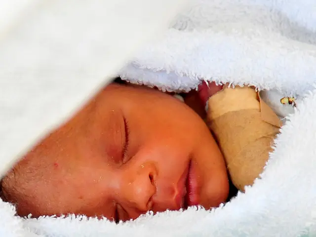 The image shows a baby sleeping peacefully in a hospital bed, wrapped in a white blanket and with a...