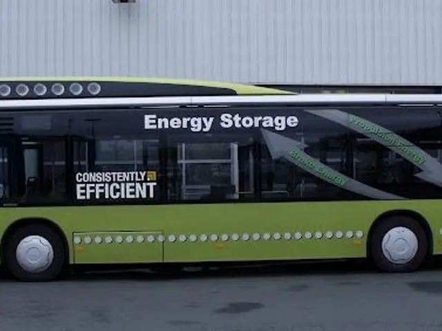 The image shows a green electric bus parked in front of a building, with a wall in the background....