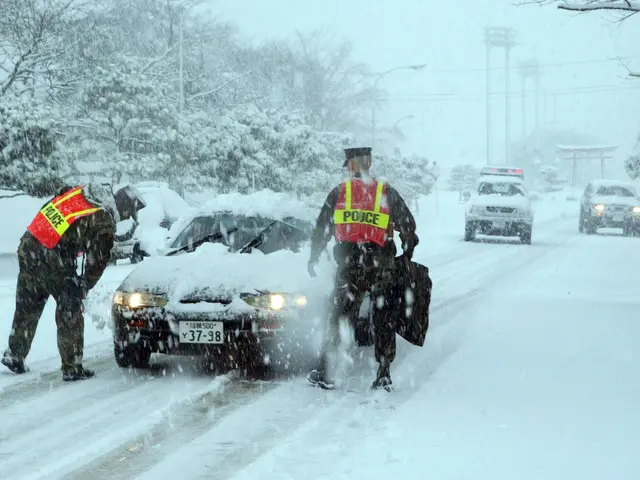 The image shows two police officers standing next to a car on a snowy road. The car is covered in a...