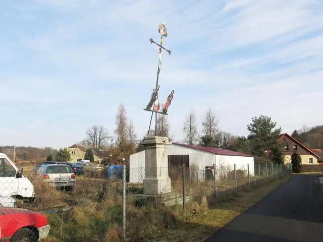 The image shows a red car parked on the side of a road next to a fence, with a cross on a pillar in...