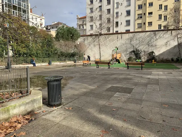 The image shows a park with benches and a trash can in the middle of it, surrounded by dried leaves...