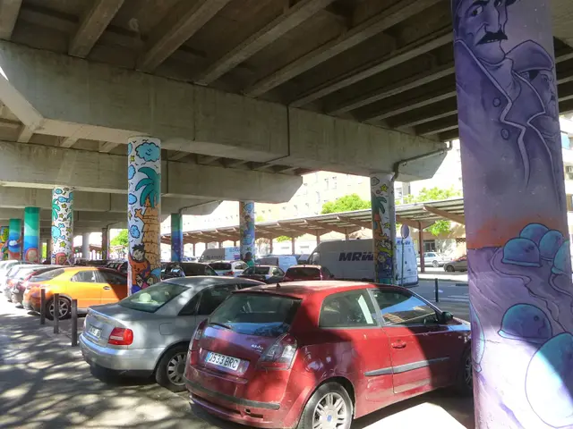 The image shows a parking lot filled with lots of parked cars under a bridge, with graffiti on the...