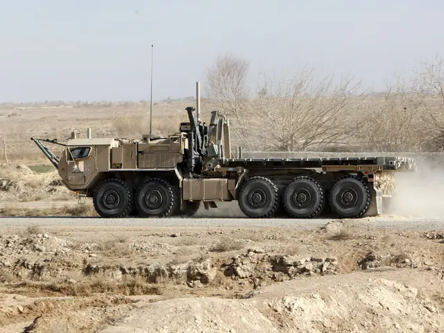 The image shows a military vehicle driving down a dirt road, surrounded by trees and a clear blue...