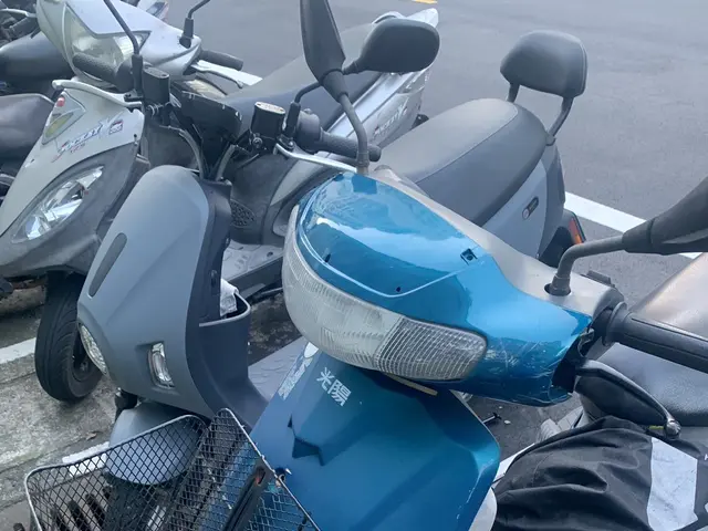 The image shows a blue scooter parked in a parking lot next to other motorcycles. The scooter is on...