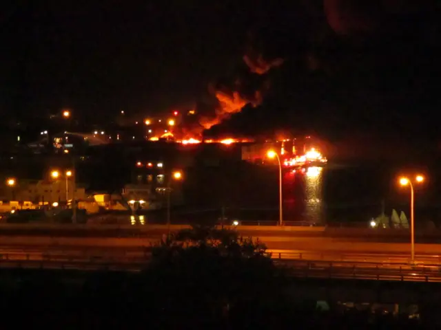 The image shows a large fire burning in the middle of a city at night, with street lights...