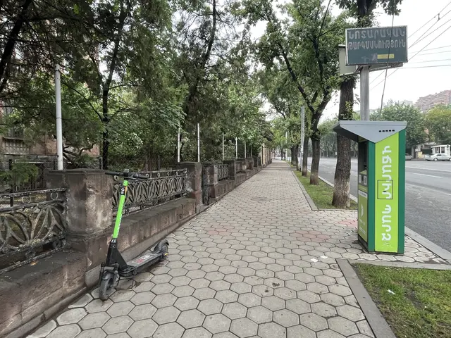 The image shows an electric scooter parked on the side of a road next to a bus stop. On the right...