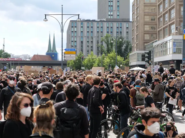 The image shows a large group of people wearing masks walking down a street lined with trees, light...