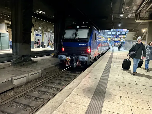 The image shows a train pulling into a train station at night. There are people walking on the...