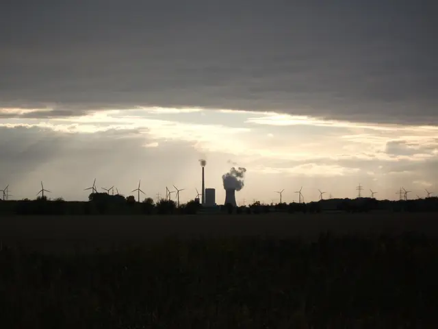 The image shows a power plant with wind turbines in the background, surrounded by trees and plants...