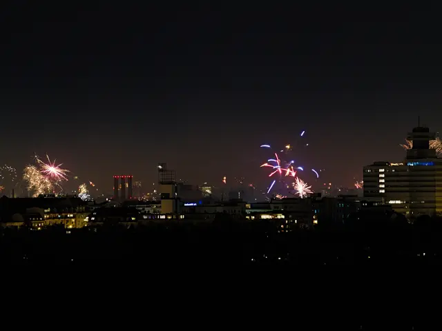 The image shows a city skyline illuminated by fireworks in the night sky. The buildings are lit up...