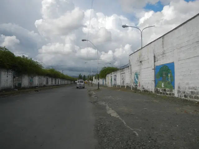 The image shows a car driving down a street lined with a wall with graffiti on it, street poles,...