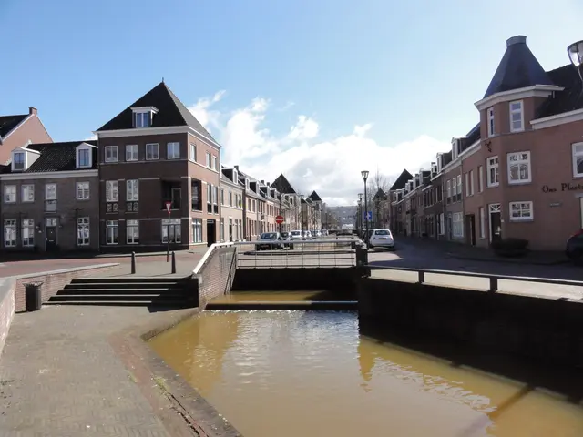The image shows a small canal running through a city street next to tall buildings, with water,...