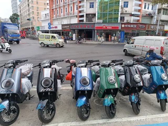 The image shows a row of electric scooters parked on the side of a street, with a person wearing a...