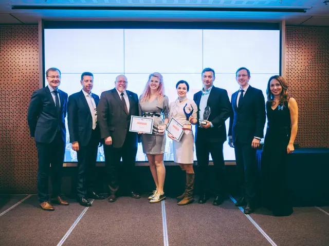 The image shows a group of people standing next to each other in front of a screen, smiling and...