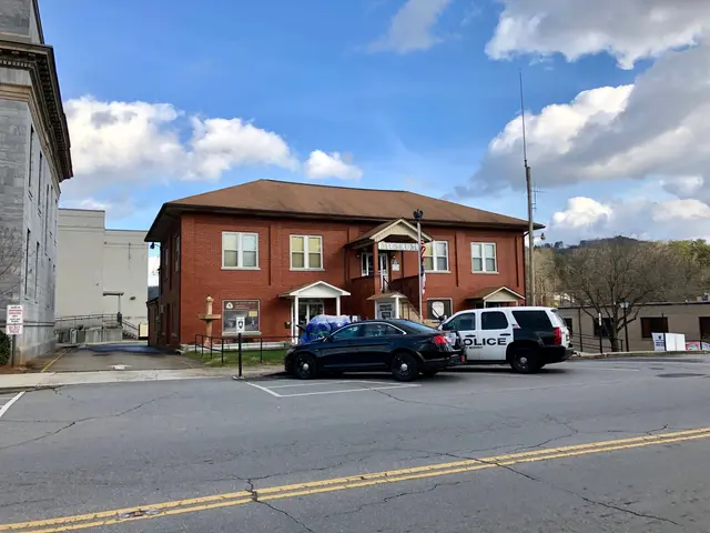 The image shows a police car parked in front of a brick building with windows, surrounded by trees,...