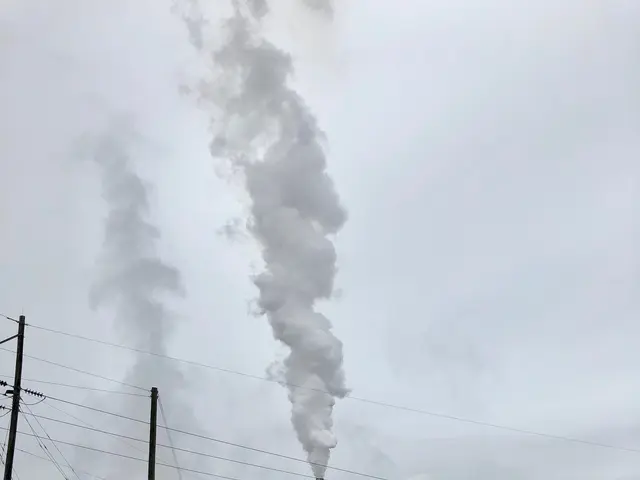 The image shows a truck driving down a road with a factory in the background, emitting smoke from...