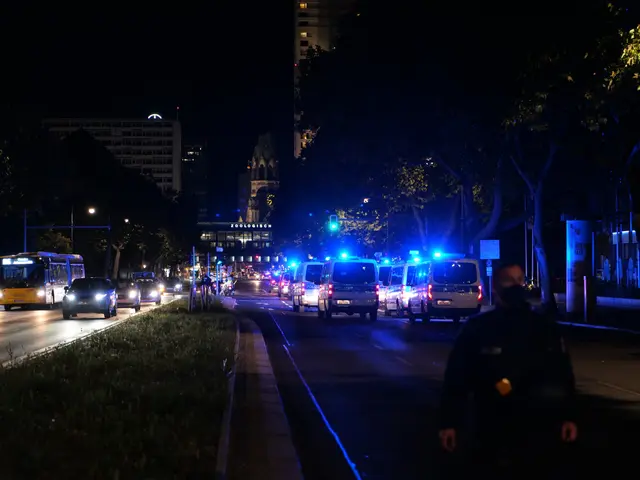 The image shows a city street at night with police cars and buses driving down it. There are people...