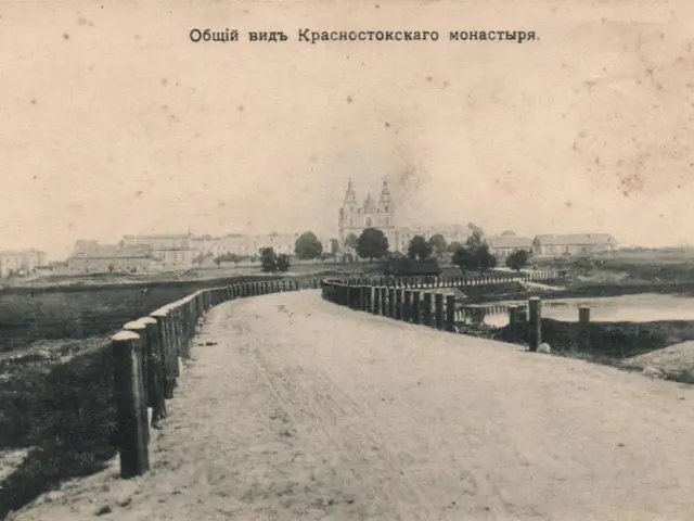 The image shows an old black and white photo of a road with a fence running along it, surrounded by...