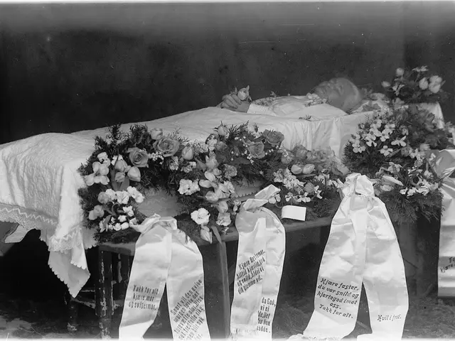 The image shows a black and white photo of a casket with flowers on it, draped with a white cloth...