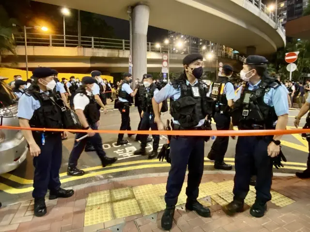 The image shows a group of police officers standing in front of a crowd of people wearing uniforms,...