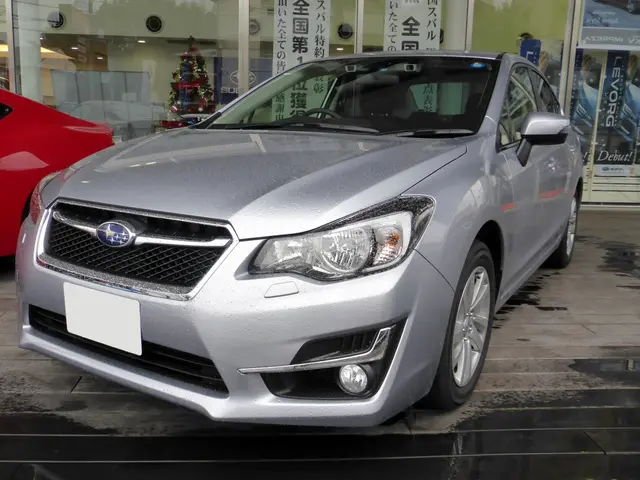 The image shows a silver car parked in front of a red car, with a glass wall in the background....