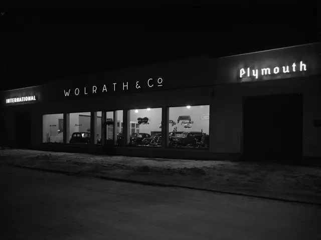 The image shows a black and white photo of a car dealership at night, with a road in the foreground...