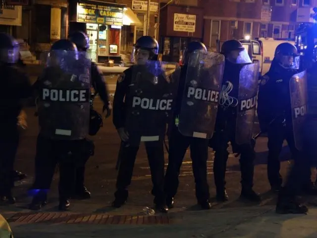 The image shows a group of police officers standing in the middle of a street at night, wearing...