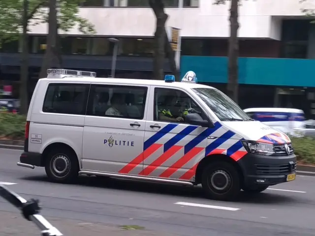 The image shows a police van driving down a street next to a bike. There are people inside the van,...