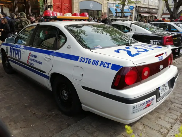 The image shows a police car parked on the side of a street, surrounded by a group of people...