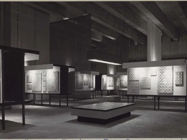 The image shows a black and white photo of a room filled with lots of tables and chairs,...