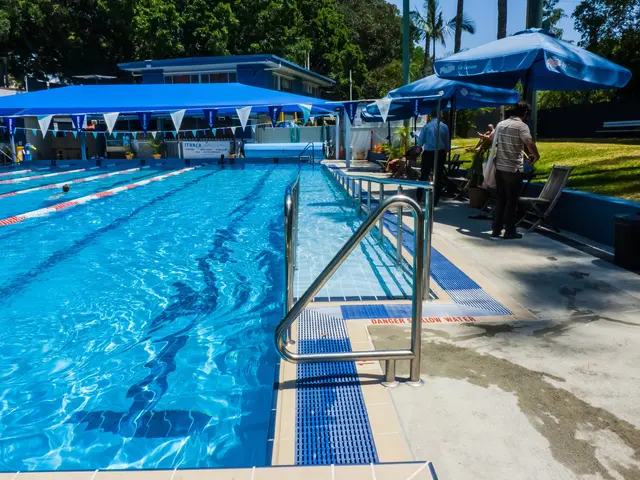 The image shows a large swimming pool with people swimming in it, surrounded by railings,...