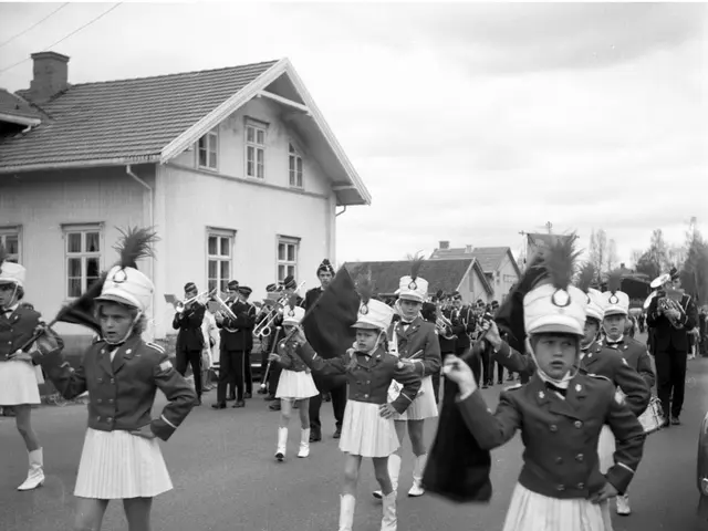 The image shows a group of young girls in uniform marching down a street, wearing caps and holding...
