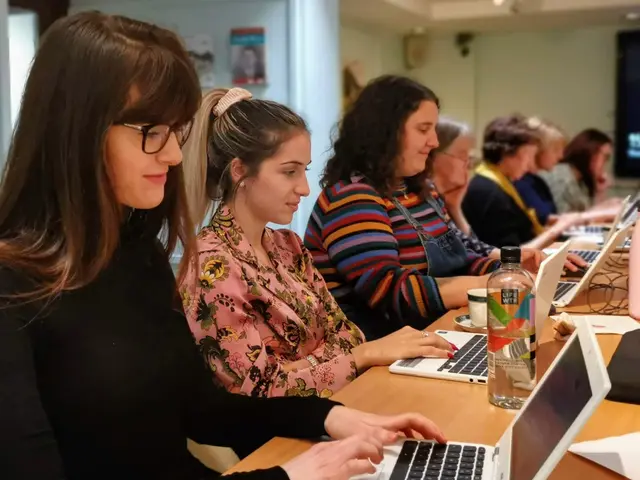 The image shows a group of women sitting around a table with laptops, bottles, cups, papers, and...