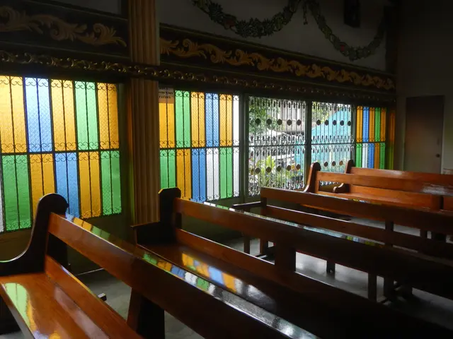 The image shows a church with wooden pews in front of a stained glass window. The window is adorned...