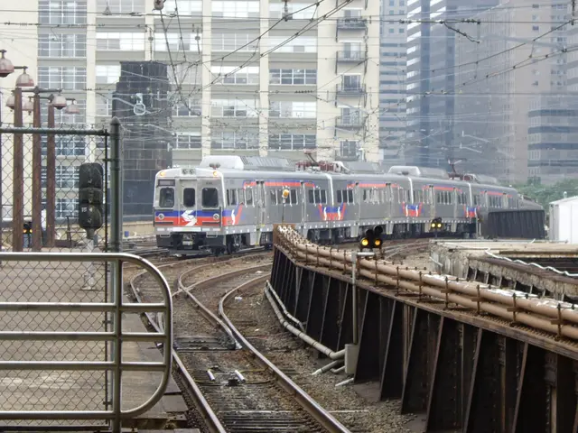 The image shows a commuter train traveling down train tracks next to tall buildings, with railway...