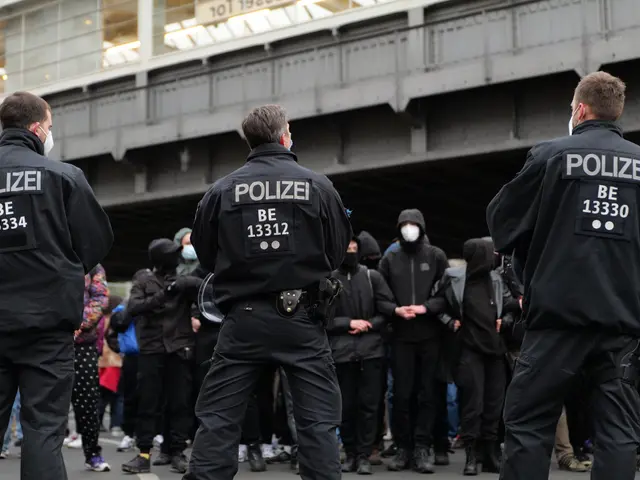 The image shows a group of police officers standing in front of a crowd of people wearing black...