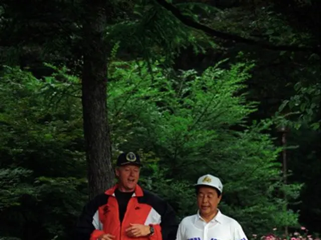 The image shows two men jogging down a path in a park, surrounded by lush green grass and tall...