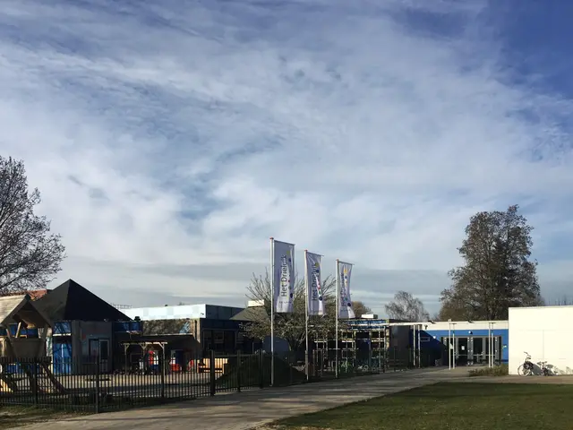 The image shows a large building with a lot of grass in front of it, surrounded by a metal fence, a...