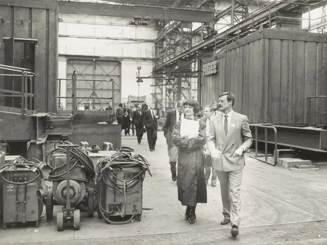 The image shows a group of people walking through a factory, with machines on the left side and a...