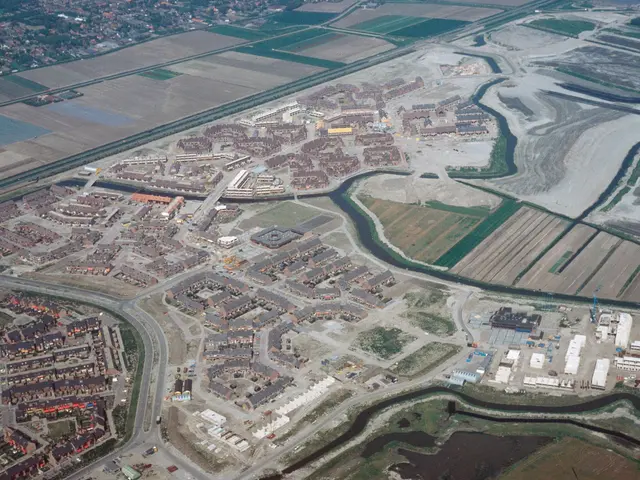 The image shows an aerial view of a city with lots of buildings, roads, fields, trees, and water....