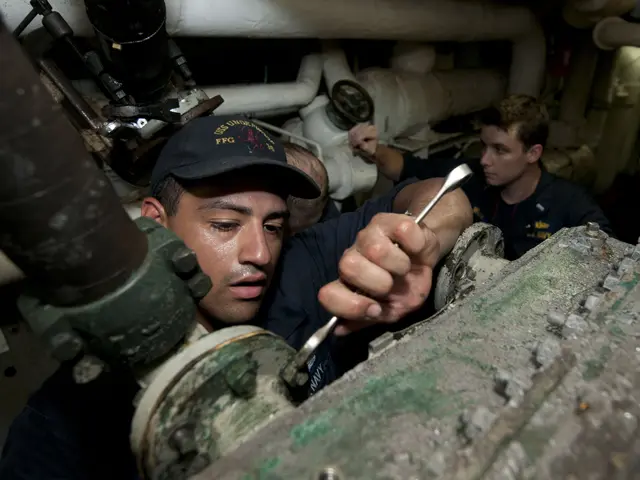 The image shows two men working on a machine in a factory, with one of them holding a tool in his...